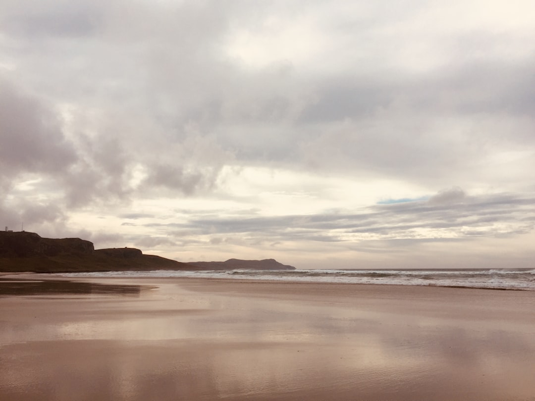 A wide, low-tide beach on the south coast of Islay. Wet sand reflects a grey-lavender sky; a rocky headland juts out on the left; distant Atlantic waves roll in from the horizon. This is the coastal environment shared by Ardbeg, Lagavulin, and Laphroaig, all sited within three miles of each other on the same southern shore.